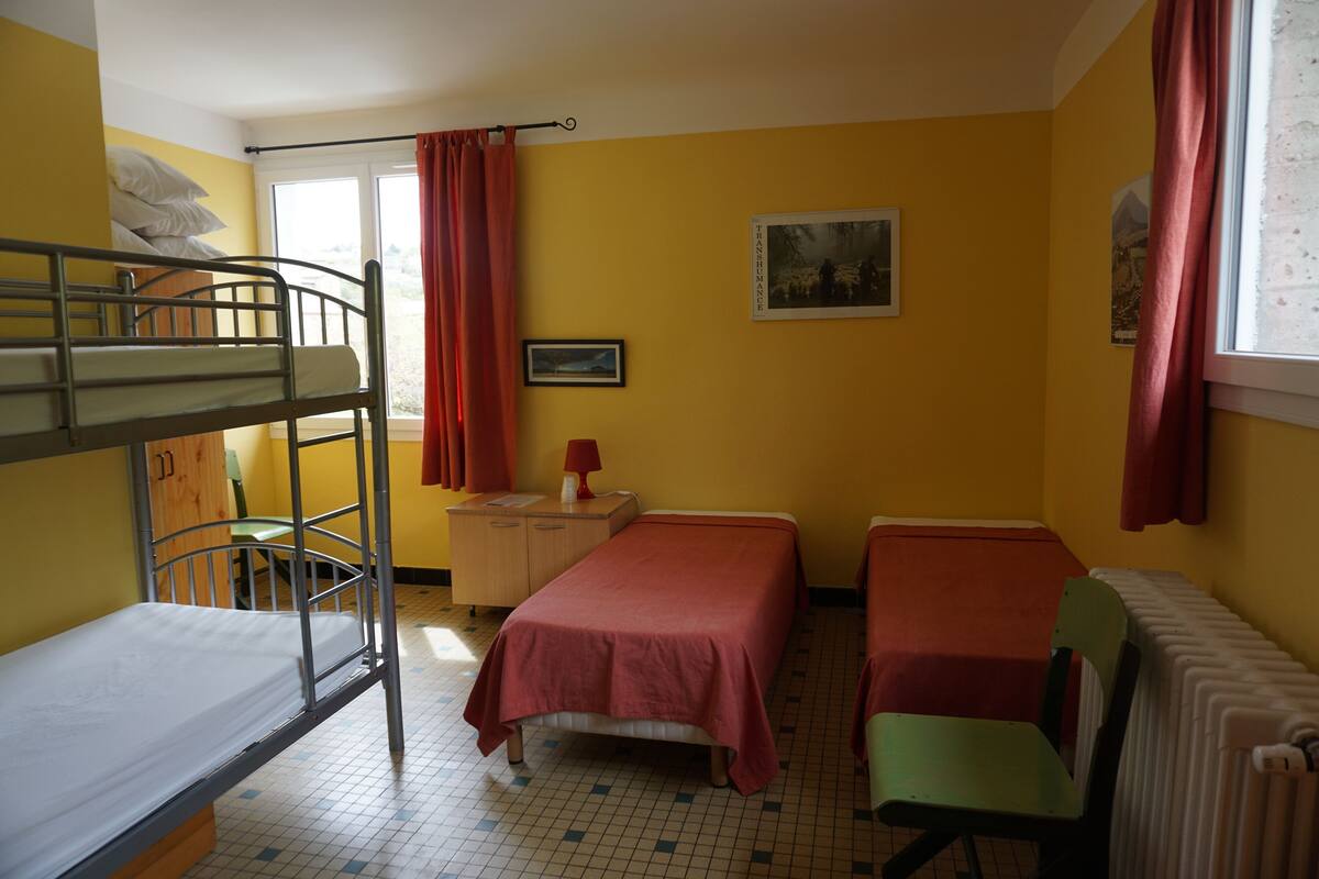 A dormitory room with bunk beds, a single bed, a desk with a chair, a radiator, and a window with curtains.