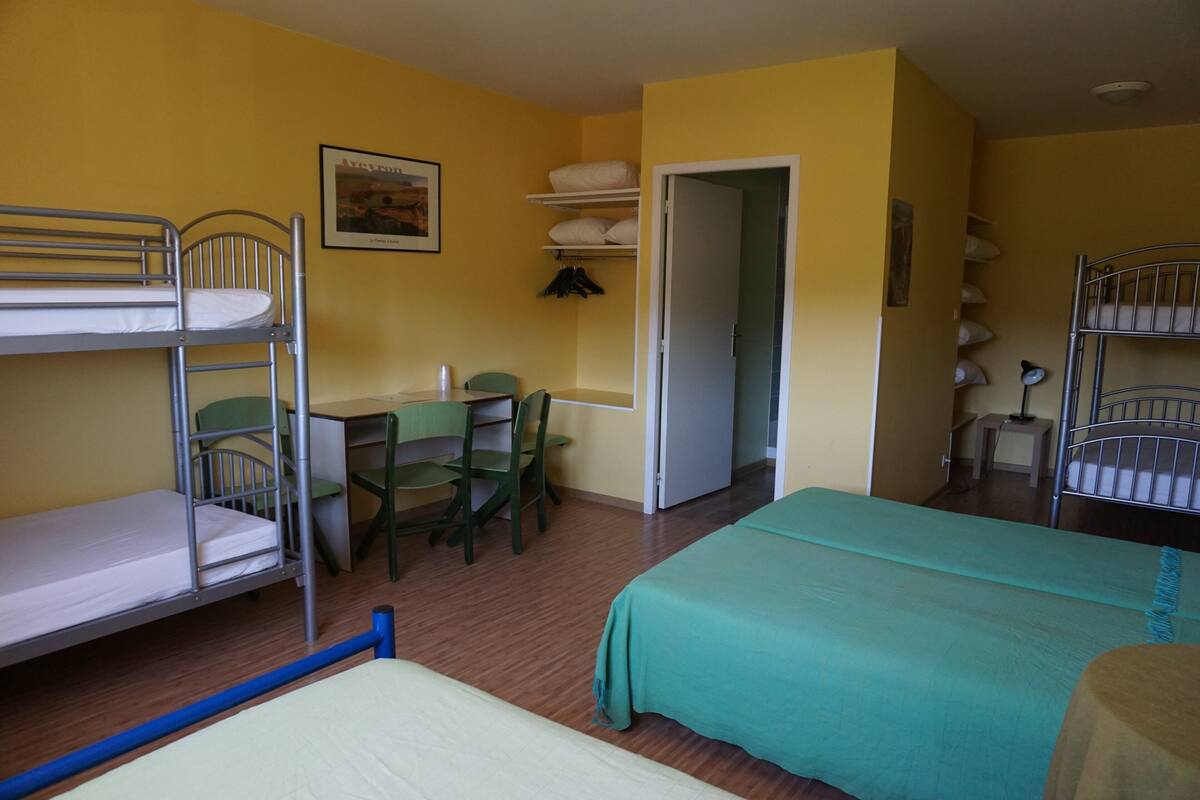 A dormitory room with bunk beds, a desk, chairs, and a bed with a green bedspread.