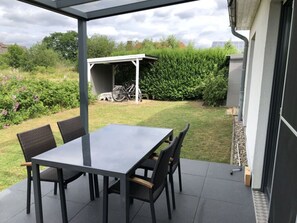 Um pátio externo com mesa e cadeiras, rodeado por um jardim e uma área coberta com uma bicicleta.