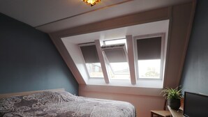 Un dormitorio con techo inclinado, una cama con colcha estampada, una mesita con una planta y un televisor sobre un soporte de madera.