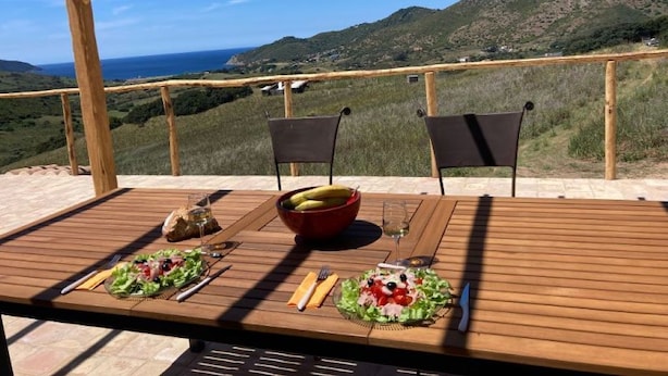 Taula de fusta a l'exterior amb dues cadires, un bol de fruita i un got de vi, davant d'una vista panoràmica d'una collada.