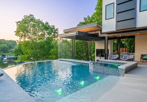 Una piscina moderna al aire libre con una cascada, rodeada de exuberante vegetación y una casa contemporánea.