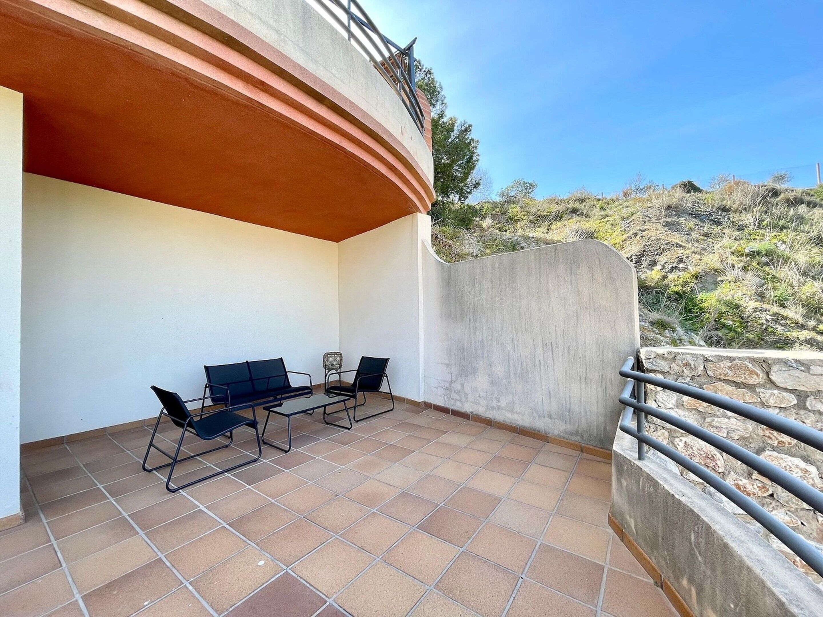 A terrace with a table and two chairs, a curved wall, and a view of a hilly landscape.