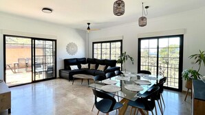 Amplia sala de estar con mesa de comedor de vidrio, sofás negros y ventanas grandes.