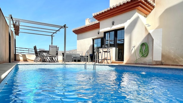 Una piscina con superficie de agua azul cristalina, rodeada por una pérgola con sillas, y un edificio con techo de tejas y puertas de cristal.