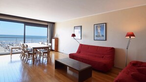 A modern living room with a red sofa, a wooden coffee table, and a dining area with chairs and a table. Large windows offer a view of the beach. - Apartment on Belgian Coast With Sea View (De Haan)