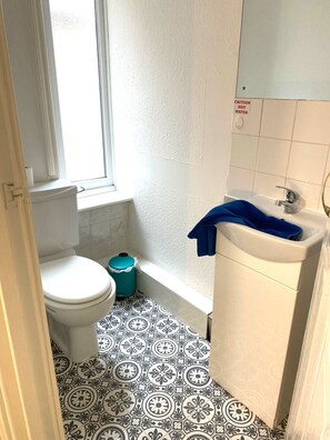 Un baño pequeño con inodoro, lavabo y bañera. Hay un letrero de precaución en la pared y una ventana sobre el inodoro.