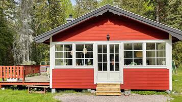 Una cabaña de madera roja con puerta y ventanas blancas, rodeada por una terraza y un área de grava, ubicada en un bosque.