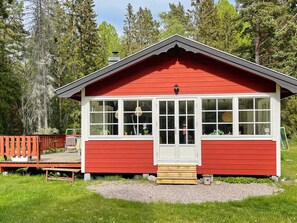 A red wooden cabin with a white door and windows, surrounded by a deck and gravel area, set in a forest. - 4 Star Holiday Home in Grisslehamn (Grisslehamn)