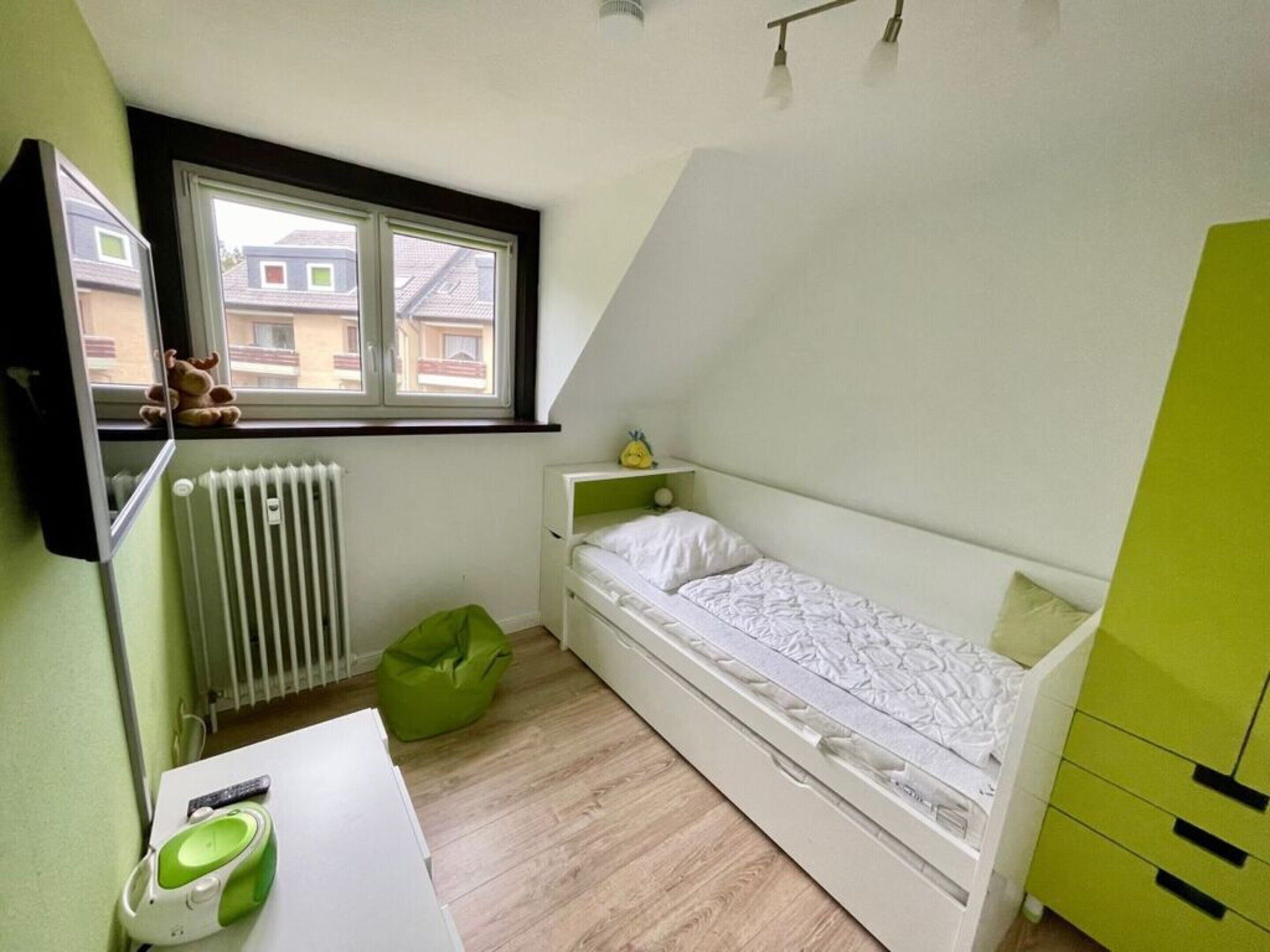 Une petite pièce bien éclairée, avec un lit simple, un radiateur, une fenêtre et un fauteuil en forme de haricot vert.