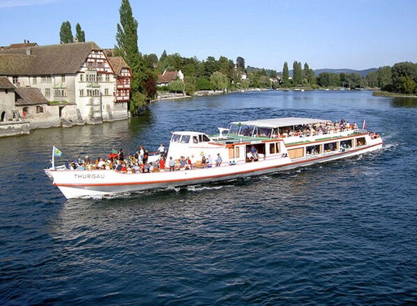 Un barco de paseo fluvial con varias cubiertas y pasajeros, rodeado de edificios y árboles a lo largo de la orilla del río.