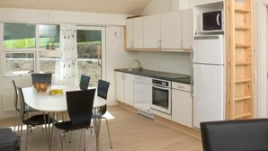 Ett kompakt kök med vita skåp, en inbyggd ugn och en mikrovågsugn. Här finns ett matbord med stolar och ett fönster med utsikt över en trädgård.