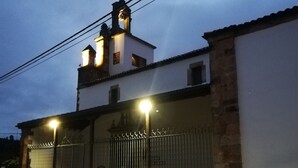 A two-story building with a white facade and a small bell tower. There are two lit windows on the upper floor and a metal gate at the entrance.