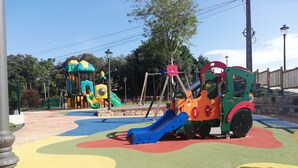 A playground with colorful play structures, including slides and climbing areas, surrounded by a paved area with a fence.