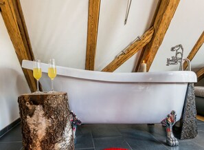 A bathtub with a wooden ceiling and a stone base, two glasses of yellow liquid on a tree stump, and a candle.