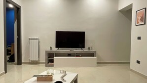 Ein modernes Wohnzimmer mit einem Flachbildfernseher auf einer weißen Unterhaltungsecke, einem kleinen Tisch mit Kerze und Kiste sowie einem grauen Teppichboden.