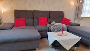 Ein modernes Wohnzimmer mit einem grauen Ecksofa, roten Kissen, einem kleinen Beistelltisch mit Kerze und einem Buch auf dem Sofa.