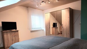 Ein Schlafzimmer mit einem Bett, einem Fernseher auf einer Holzkommode, einem großen Spiegel und einem Fenster mit transparenten Vorhängen.