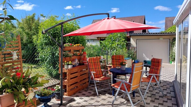 Eine Terrasse im Freien mit einem roten Sonnenschirm, Holzhöckern und einem Tisch.