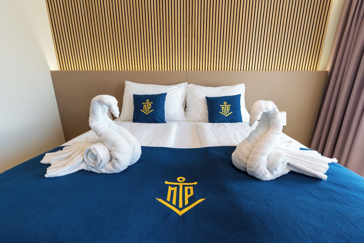 Habitación de hotel con una cama bien hecha, dos almohadas con un logotipo de escudo y una colcha azul con el mismo logotipo.