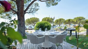 Un espace de restauration en plein air avec une table ronde et des chaises en osier blanc, entouré de verdure et sous un ciel dégagé.