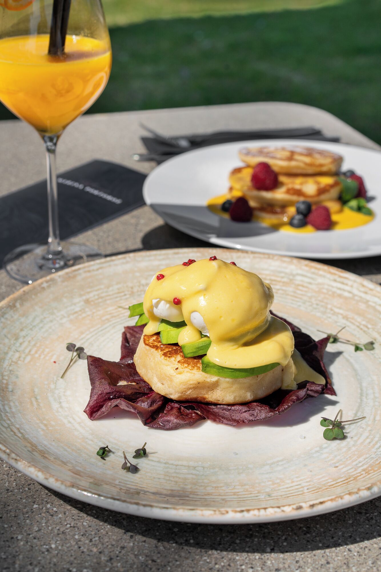 Desayuno con huevos benedictinos, un vaso de jugo de naranja y una porción de panqueques con frutos rojos.