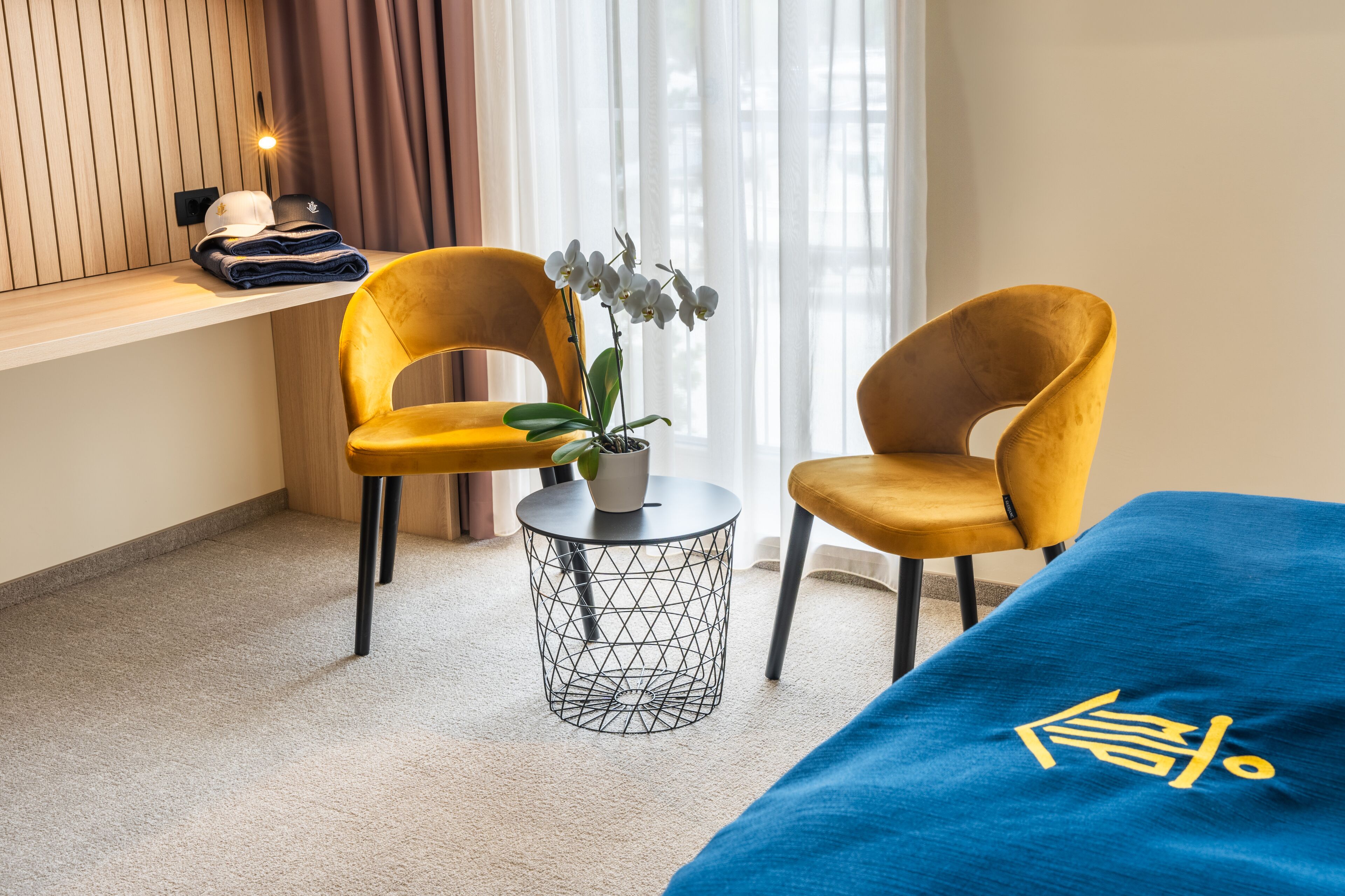 A hotel room with two yellow chairs, a small round table with a plant, a bed with a blue cover, and a shelf with folded clothes and a cap.
