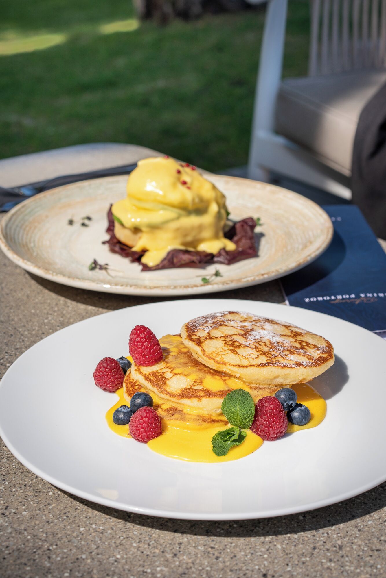 Two plates of food, one with pancakes and berries and the other with a dessert.