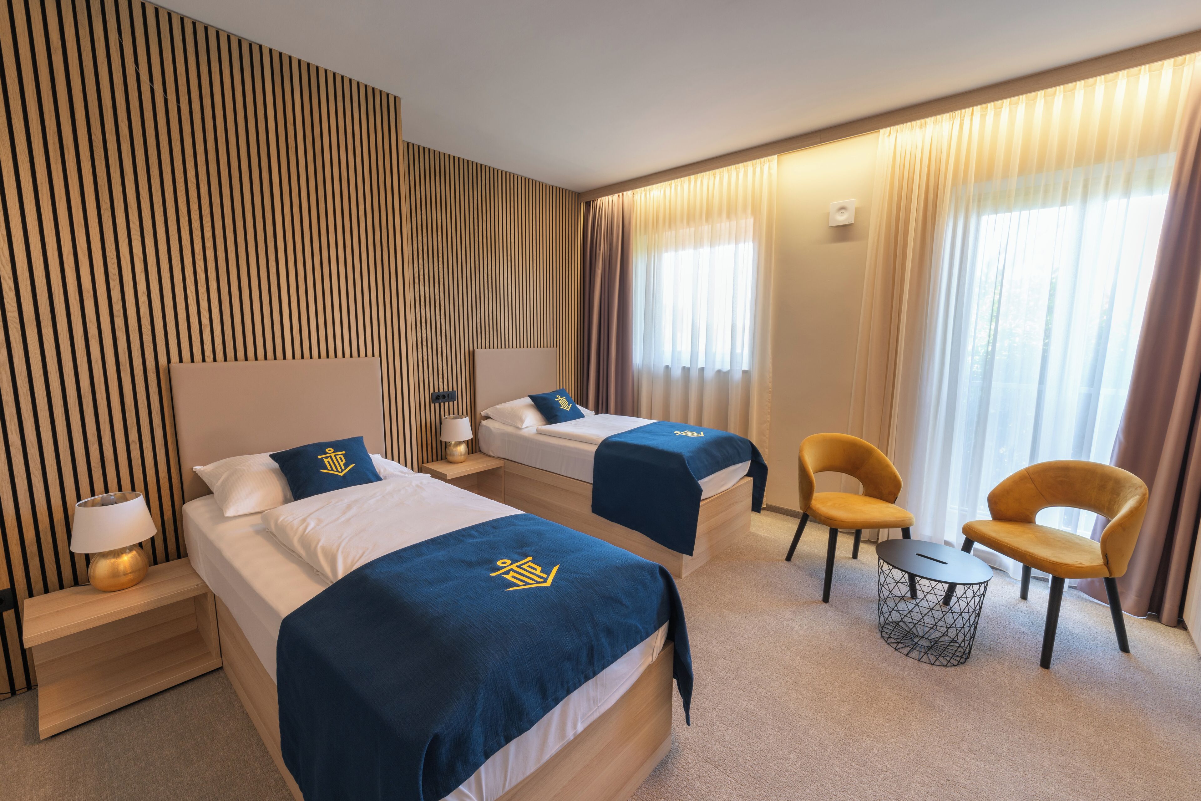 Ein Hotelzimmer mit zwei Betten, einem kleinen runden Tisch, zwei Stühlen, einer Lampe und einem Fenster mit Vorhängen.