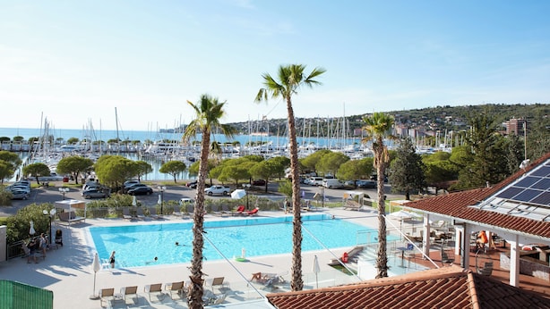 Une piscine entourée de chaises longues et de palmiers, offrant une vue sur une marina avec des bateaux et une ville côtière.
