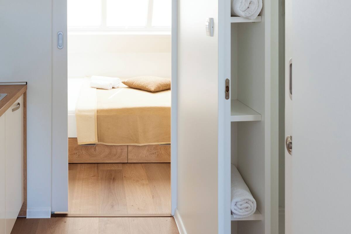 Un pasillo estrecho que conduce a un dormitorio con una cama, techo de madera y un armario blanco.