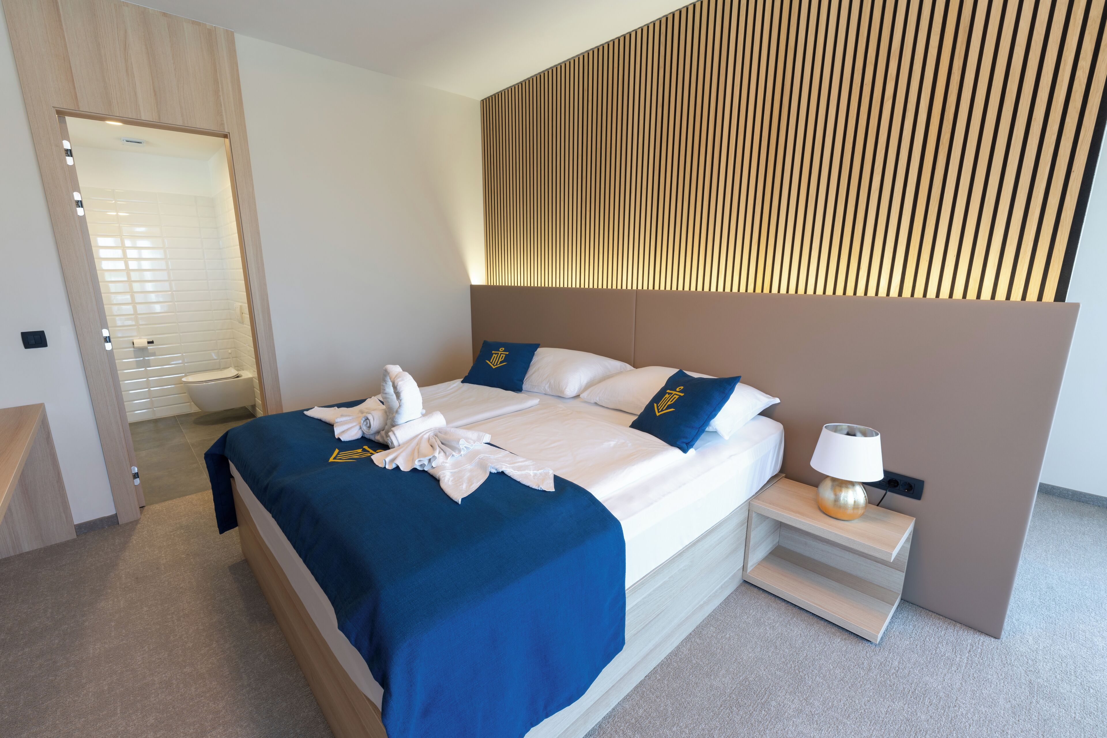 Quarto de hotel moderno com uma cama grande, mesa de cabeceira com luminária e cabeceira de ripas de madeira.