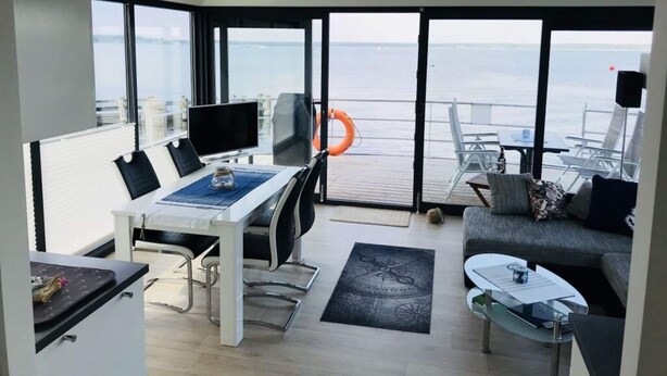 A modern living room with a large window overlooking the sea, a dining area with a table and chairs, and a cozy seating area with a sofa and coffee table. - Apartment in Mecklenburg Western Pomerania (Goehren-Lebbin)