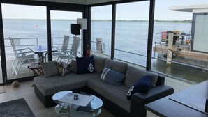 A modern living room with a large window overlooking a body of water, a sofa with cushions, a glass coffee table, and a small table with chairs. - Apartment in Mecklenburg Western Pomerania (Goehren-Lebbin)