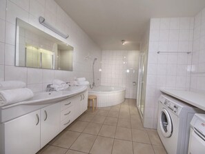 A modern bathroom with a large mirror, a sink with two basins, a bathtub, a shower area, and a washing machine. - Apartment Alpenresort Saalbach E2 (Saalbach-Hinterglemm)