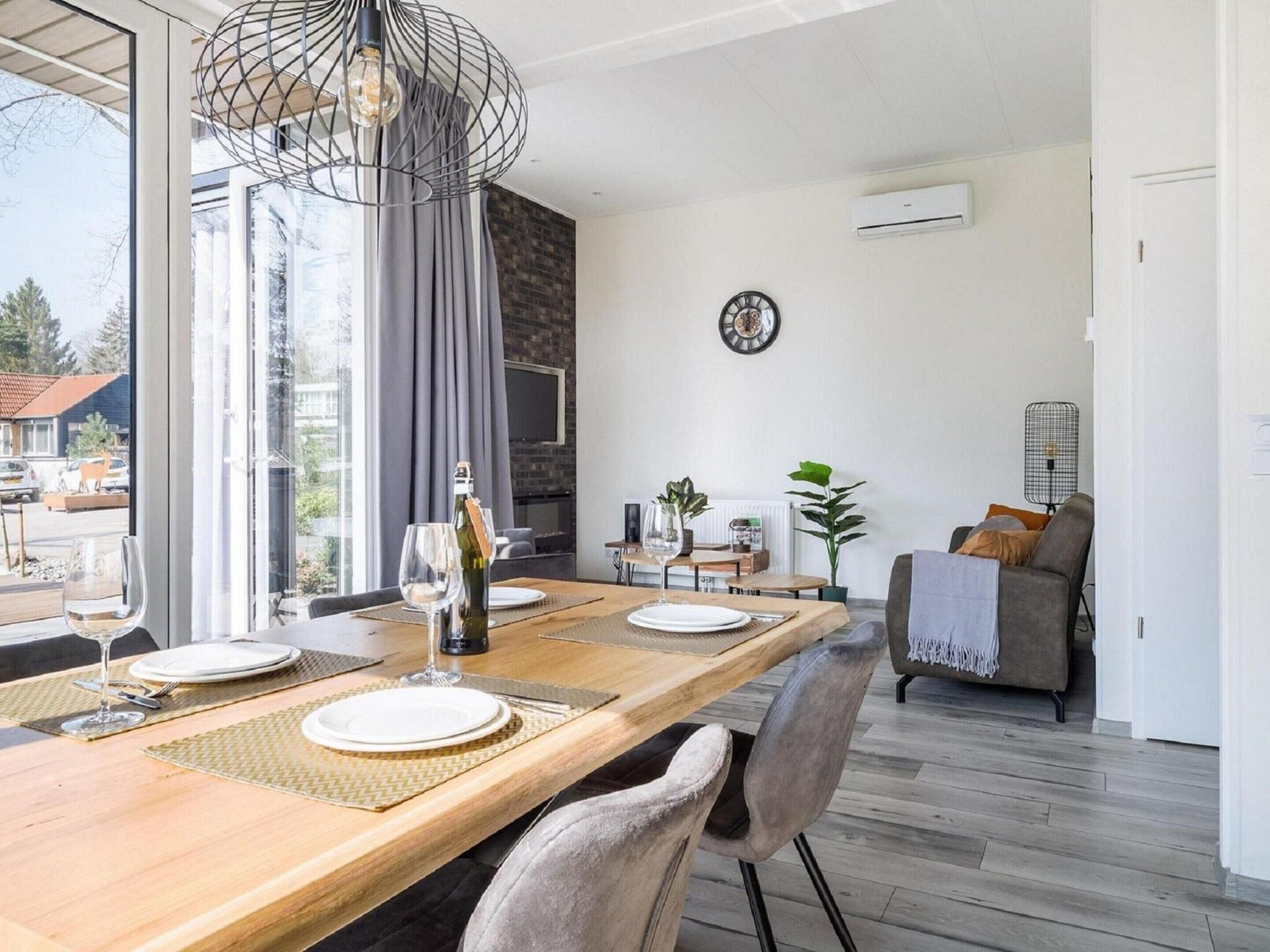 Un coin repas moderne avec une table en bois dressée pour deux personnes, une bouteille de vin et un verre posés sur la table. On y trouve une grande fenêtre avec des rideaux, un mur en briques et une horloge au mur.