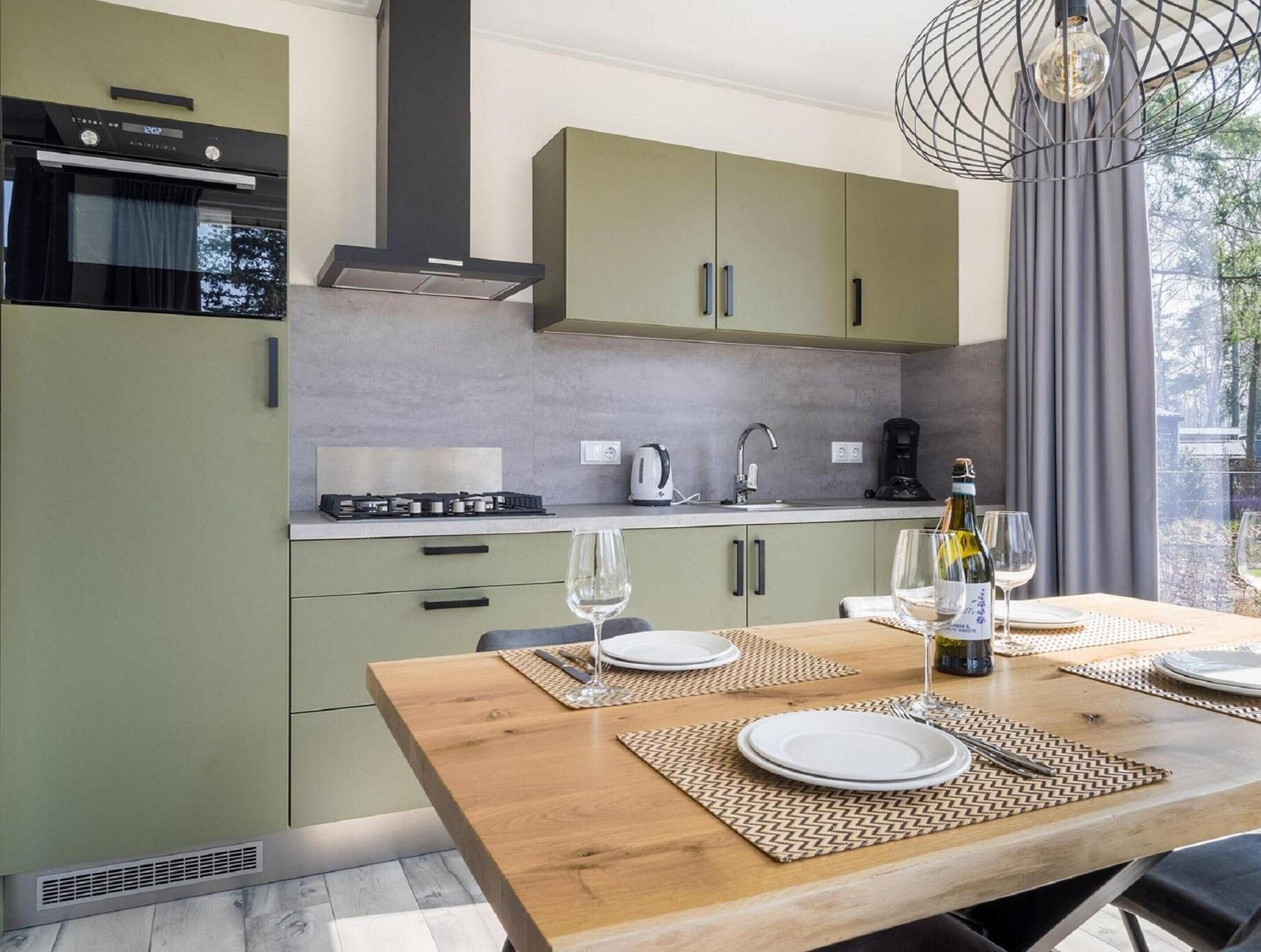 Una cocina moderna con gabinetes verdes, una mesa de comedor de madera dispuesta para dos personas, y vistas al exterior a través de una ventana.