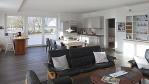 A spacious living area with a black leather sofa, a wooden coffee table, and a dining table with chairs. A kitchen is visible in the background with white cabinets and a large window. - Loft Classiccar Modern Retreat (Gluecksburg)