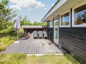 Una terraza de madera con mobiliario exterior, un espacio de estar cubierto y un amplio ventanal.