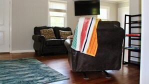 A living room with a dark leather sofa, a colorful blanket, a flat-screen TV, and a bookshelf. - Mema and Pa’s Place (Byrdstown)