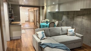 Ein modernes Wohnzimmer mit einem grauen Ecksofa, Holzböden und einem Essbereich mit blauer Tischdecke.