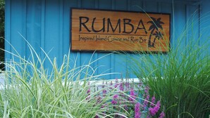 Ein Holzschild mit dem Schriftzug 'RUMBA Inspired Island Cuisine and Rum Bar' vor einer blauen Wand.