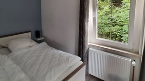 Ein Schlafzimmer mit Bett, Nachttisch, Heizkörper und einem Fenster mit Blick auf Grünflächen.