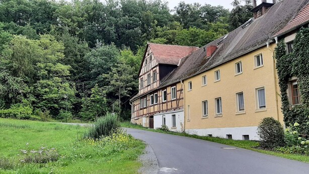 Ein traditionelles Fachwerkhäuschen mit geneigtem Dach, neben einer gepflasterten Straße, umgeben von üppigem Grün und Bäumen.