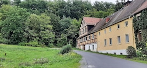 A traditional half-timbered house with a sloped roof, adjacent to a paved road, surrounded by lush greenery and trees.