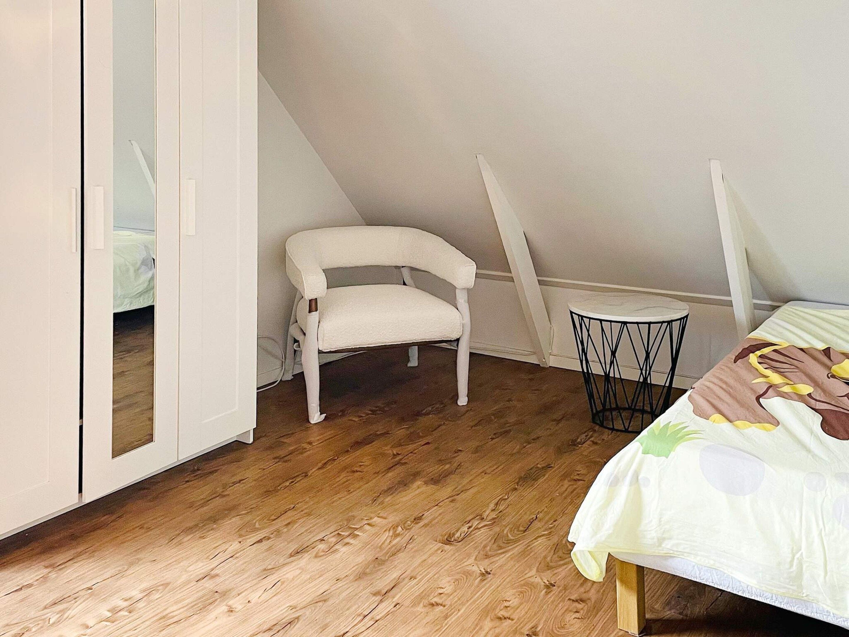 A bedroom with a sloped ceiling, a white chair, a round table, and a bed with a floral bedspread.