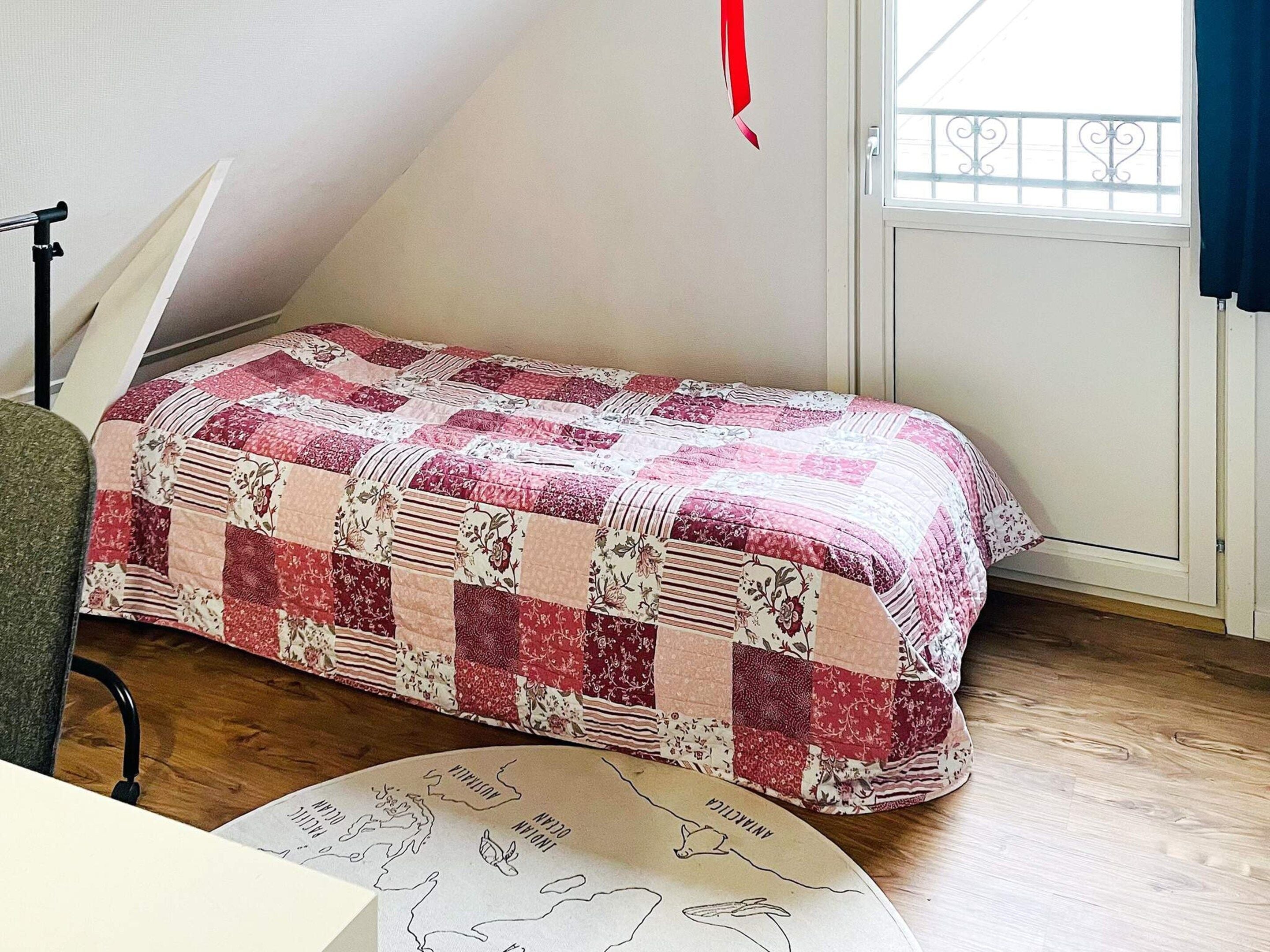 A room with a bed covered in a patchwork quilt, a desk with a chair, a window with a view, and a map on the floor.