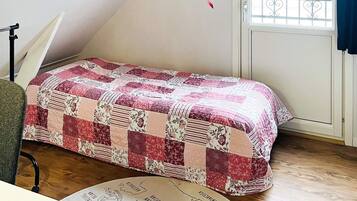 A room with a bed covered in a patchwork quilt, a desk with a chair, a window with a view, and a map on the floor.
