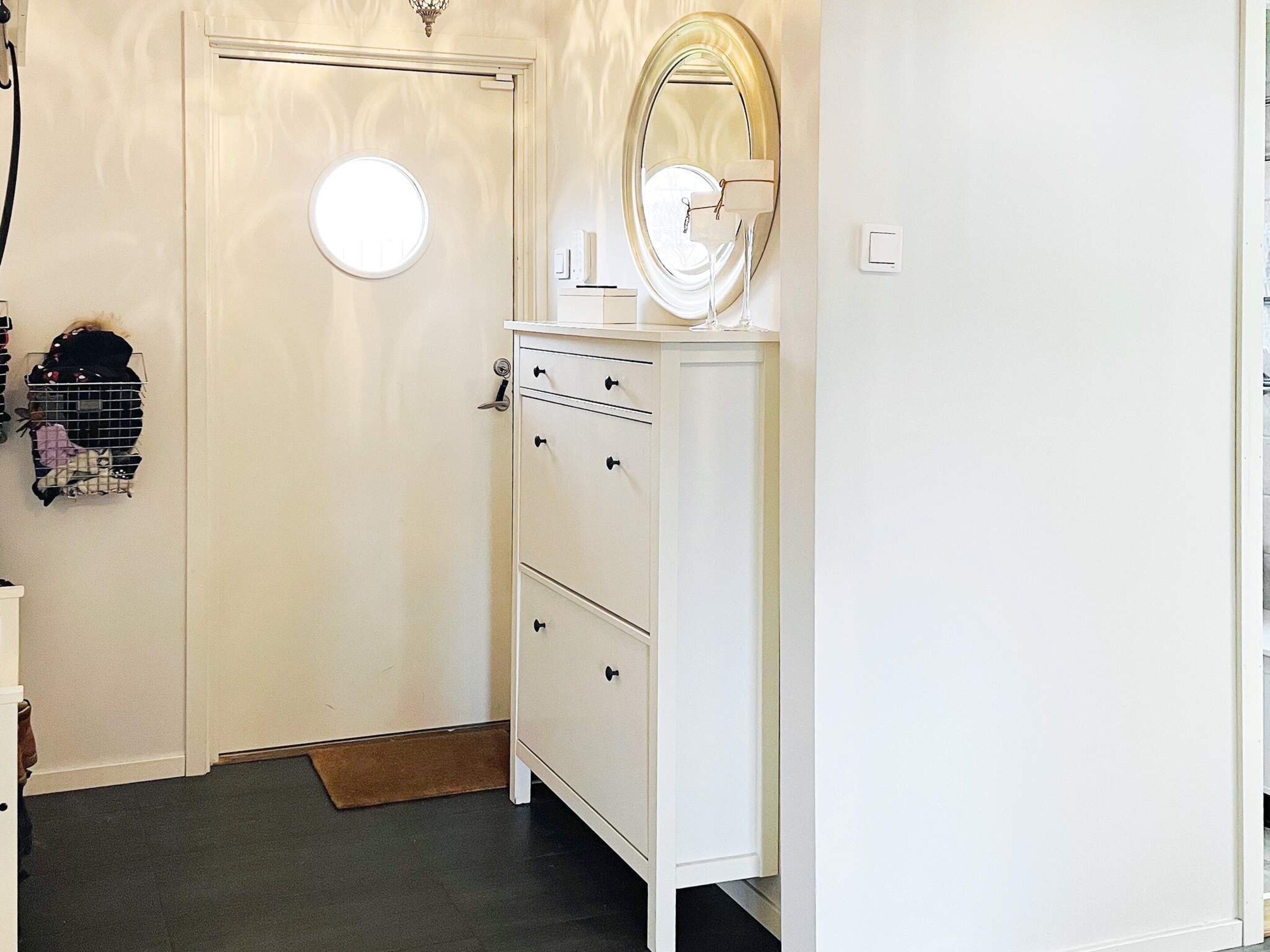 A white door with a round mirror above it, a white dresser with drawers, and a small shelf with items on the wall.