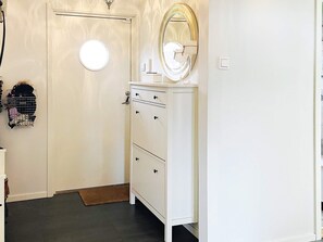 Una puerta blanca con un espejo redondo encima, un aparador blanco con cajones, y una pequeña estantería con objetos en la pared.